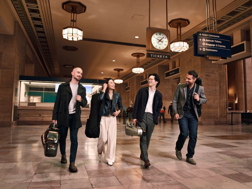 Four diverse musicians wearing casual contemporary clothing carry their string instruments in cases through the marbled lobby of a train station.