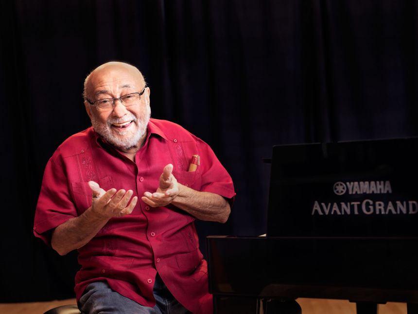 An elder gentleman wearing glasses smiles and sits at a grand piano.