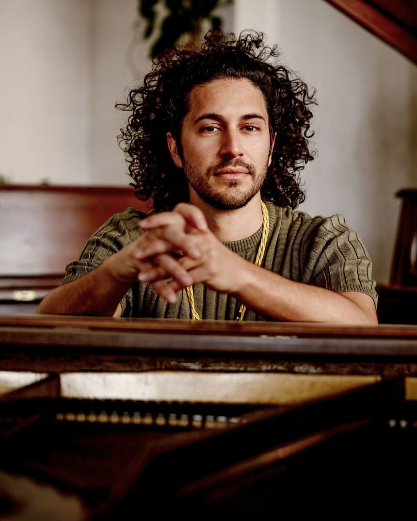 A man with his curly hair held with a headband sits with his hands folded on a piano.