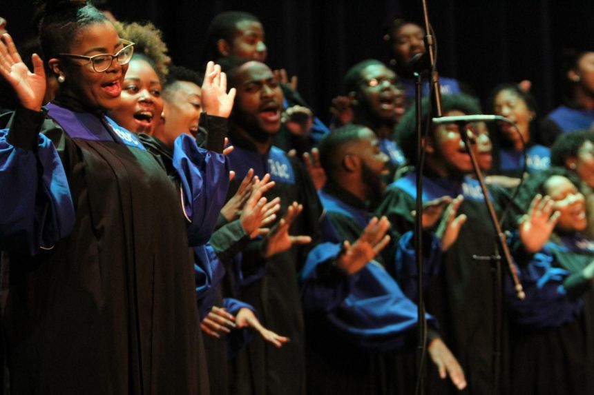 A group of African-American singers wearing matching robes stand in rows and raise their hands in song.