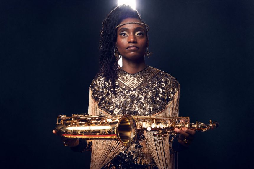 A Black woman wearing a shiny headband presents a saxophone like a treasure.