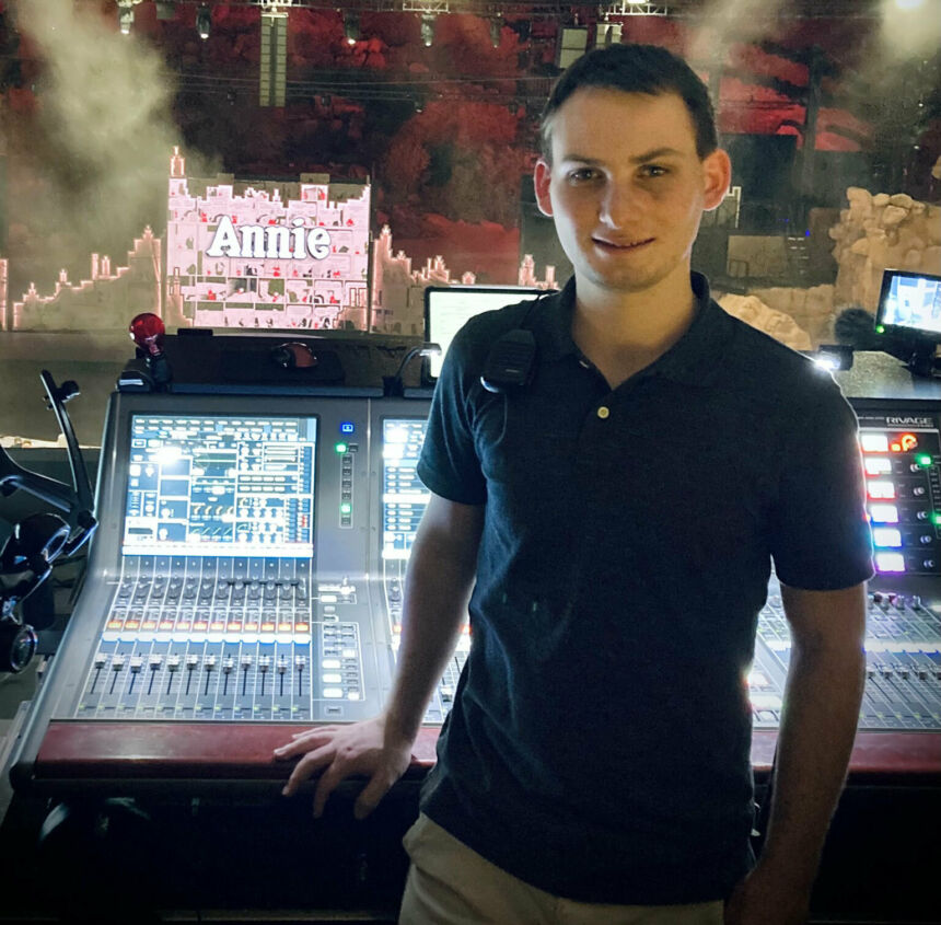 A young man stands in front of a sound production board with its knobs and buttons.