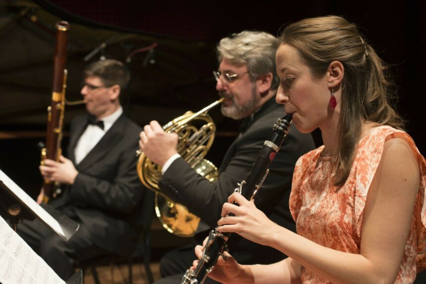 Three musicians play their wind instruments while sitting in front of their music stands.