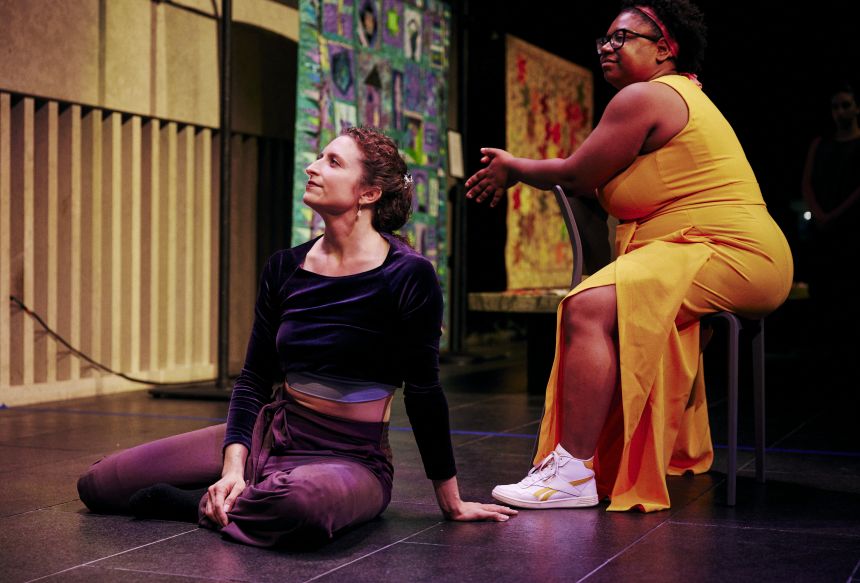 A white woman sits on a stage while a Black women sits in a chair next to her.