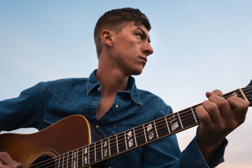 A young white man holds an acoustic guitar as he looks off into the distance.