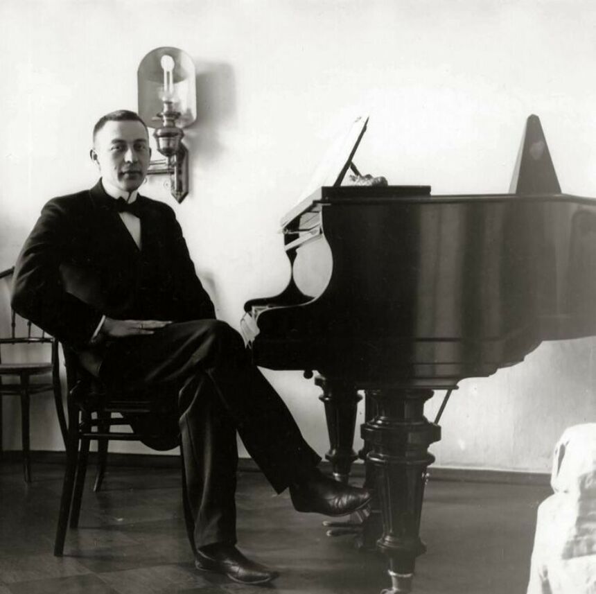 An historic photograph shows a man wearing a suit and bowtie and sitting at the keys of a piano.