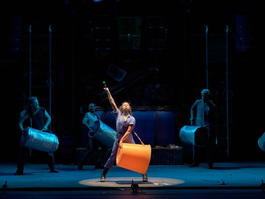 A woman of color standing center stage wears a plastic drum on a strap around her neck and shoulders as she points her arms upward.