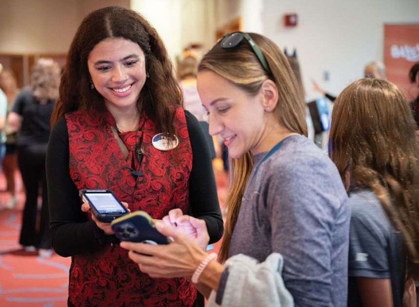 A young woman assists another woman with accessing tickets on her mobile phone.