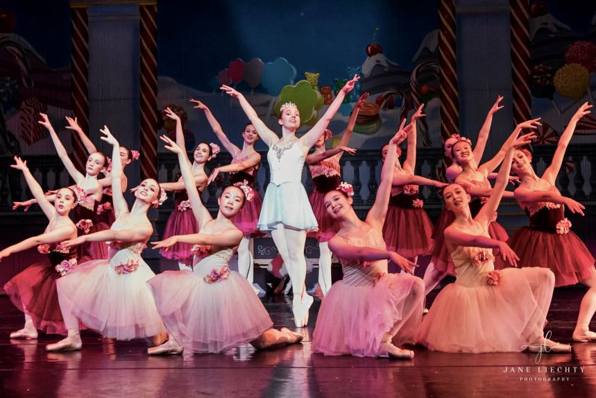 A group of young ballet dancers wear matching tutus and crowd around a dancer in the center in the pointe position.