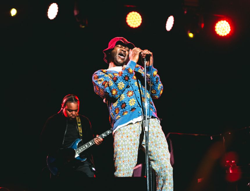 A black man wearing a crocheted suit sings passionately into a microphone on a darkened stage.