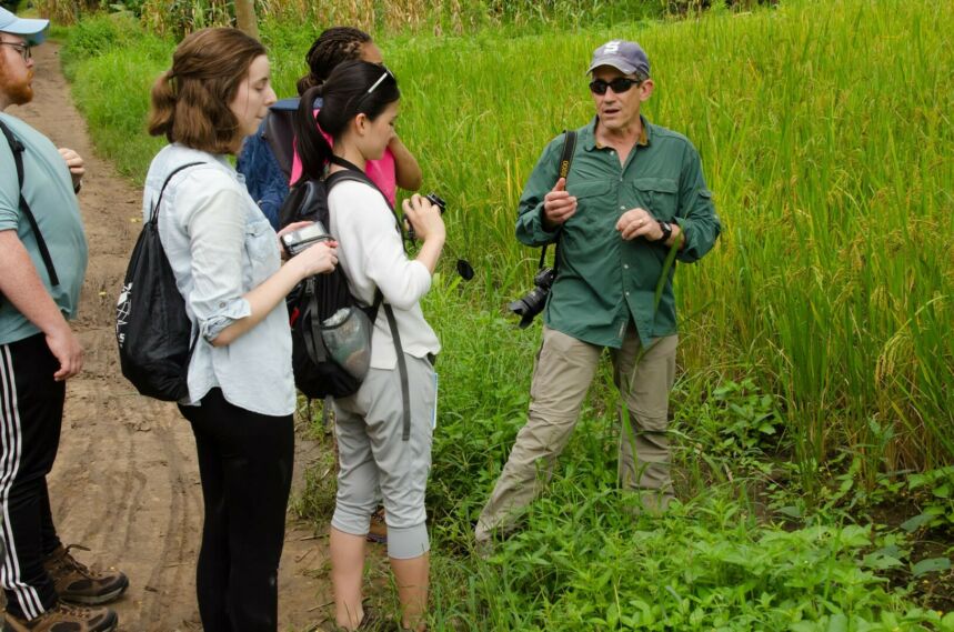Students standing on a dirt road flanking a lush field of head-high green grasses, listening to professor Larry Gorenflo.