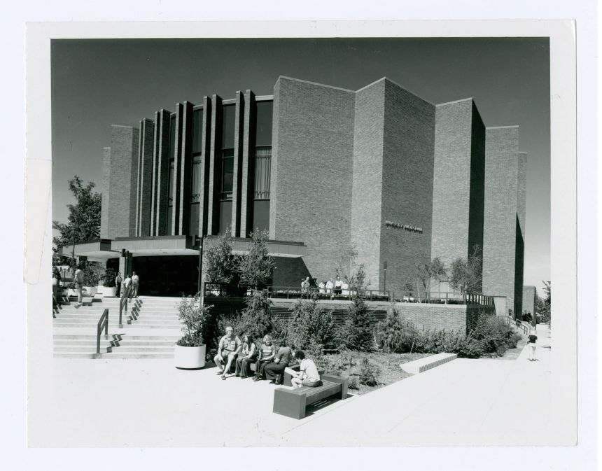 An archival photo shows the large, brick Eisenhower Auditorium and its stepped patio, landscaping and tall vertical windows.
