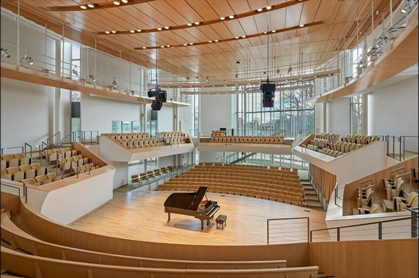 Circular concert hall with wooden stage, many windows and seats with a grand piano on stage