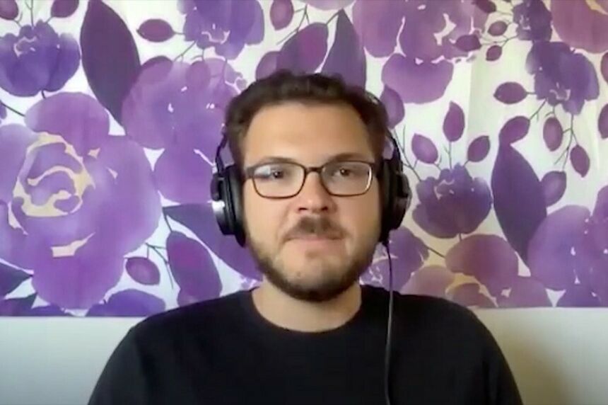 Screen shot of a man with dark hair, a beard and dark-rimmed glasses in front of a purple and white background