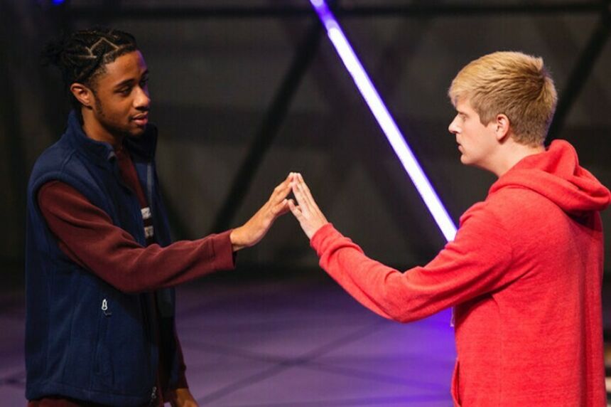 A Black young man and a white young man stand face to face touching hands