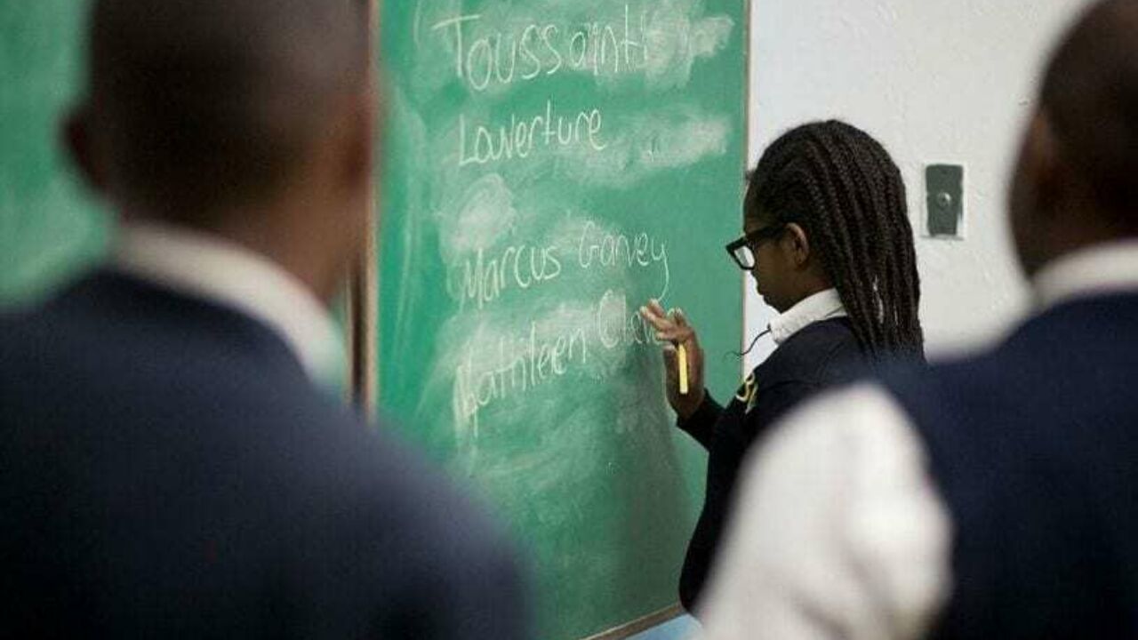 Three African American students in a classroom, with one of said students writing on a chalkboard.