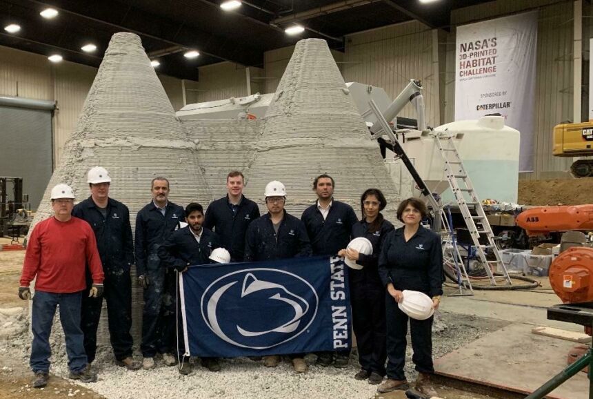 Penn State NASA Mars Challenge team standing together in front of full-size 3d-printed concrete dwellings holding Penn State flag