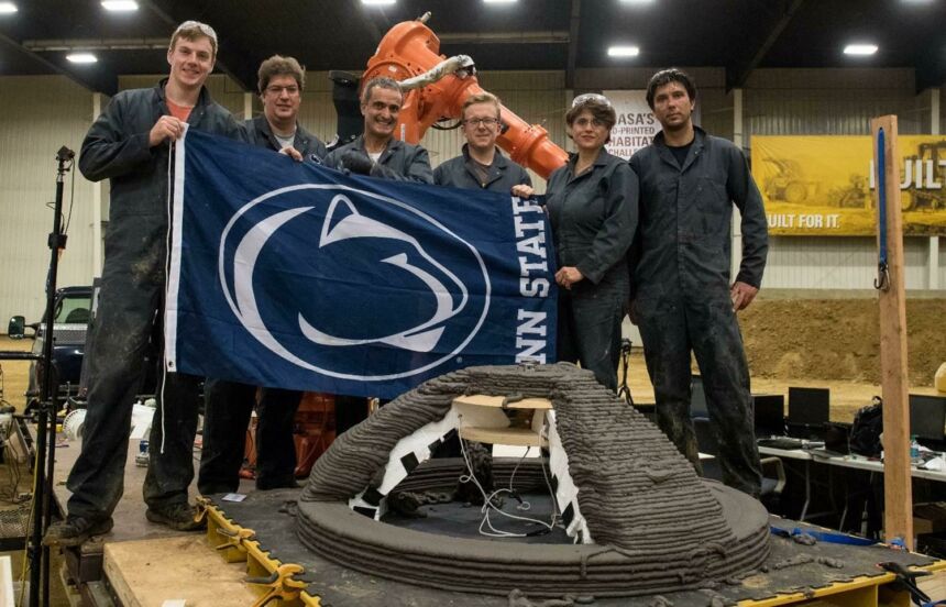 Penn State NASA Mars Challenge team standing together in front of large robotic arm, holding Penn State flag
