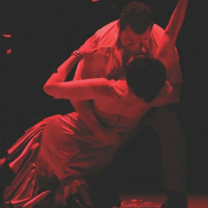 Two operatic performers dancing on a darkened, red-cast stage.