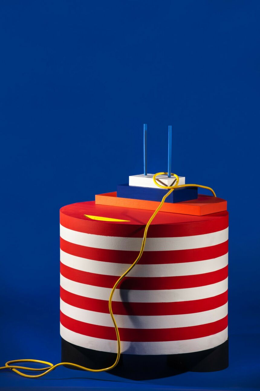 A blue background with a white and red cylinder on top of a black base. Orange, blue and white rectangles are stacked on top with two blue poles sticking out of the top of the structure. A yellow rope is hanging down from one of the poles.