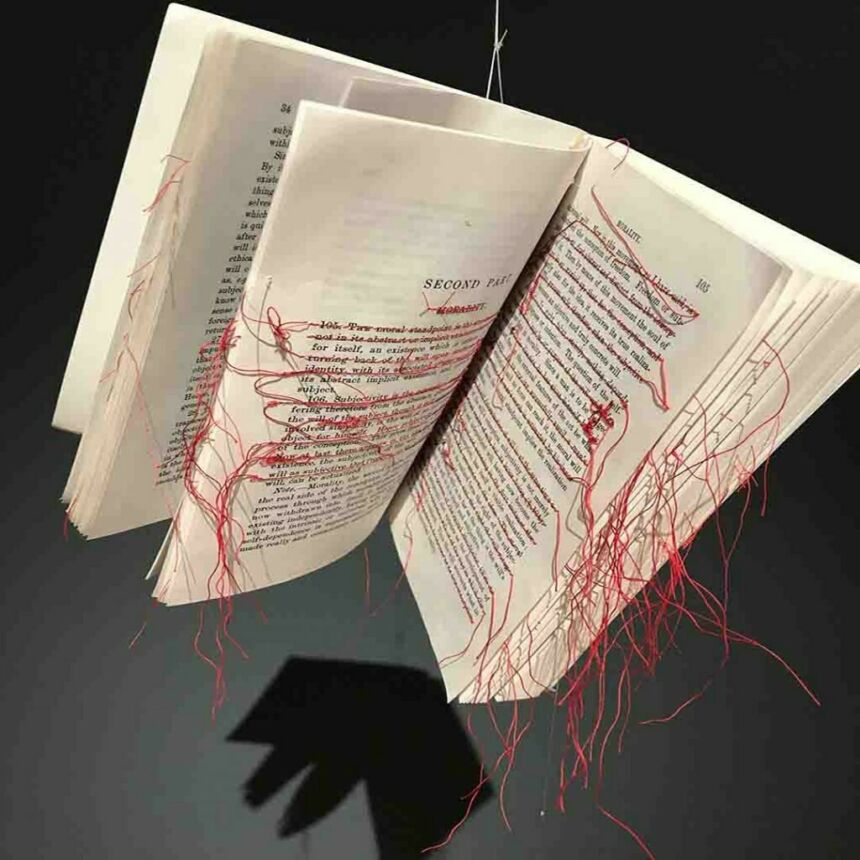 Opened book, suspended by string, with red threading stitched across various lines of text on each page.