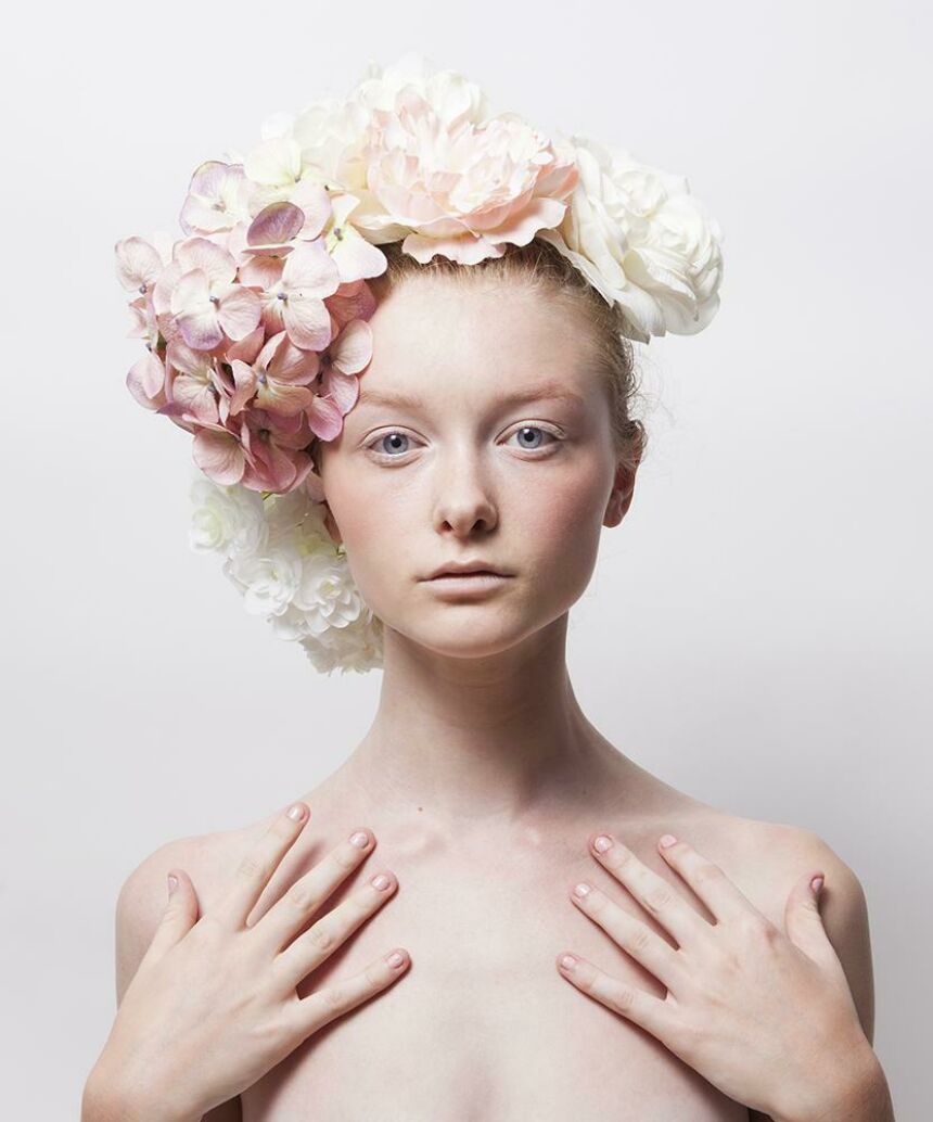 Studio headshot of a young woman wearing a variety of pastel spring flowers in her hair, her hands placed against the front of her shoulders.