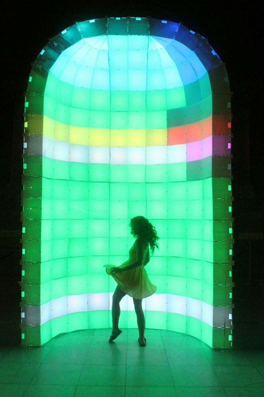 Silhouette of a young woman in short dress dancing in front of a tall, rounded alcove made of rainbow light boxes.