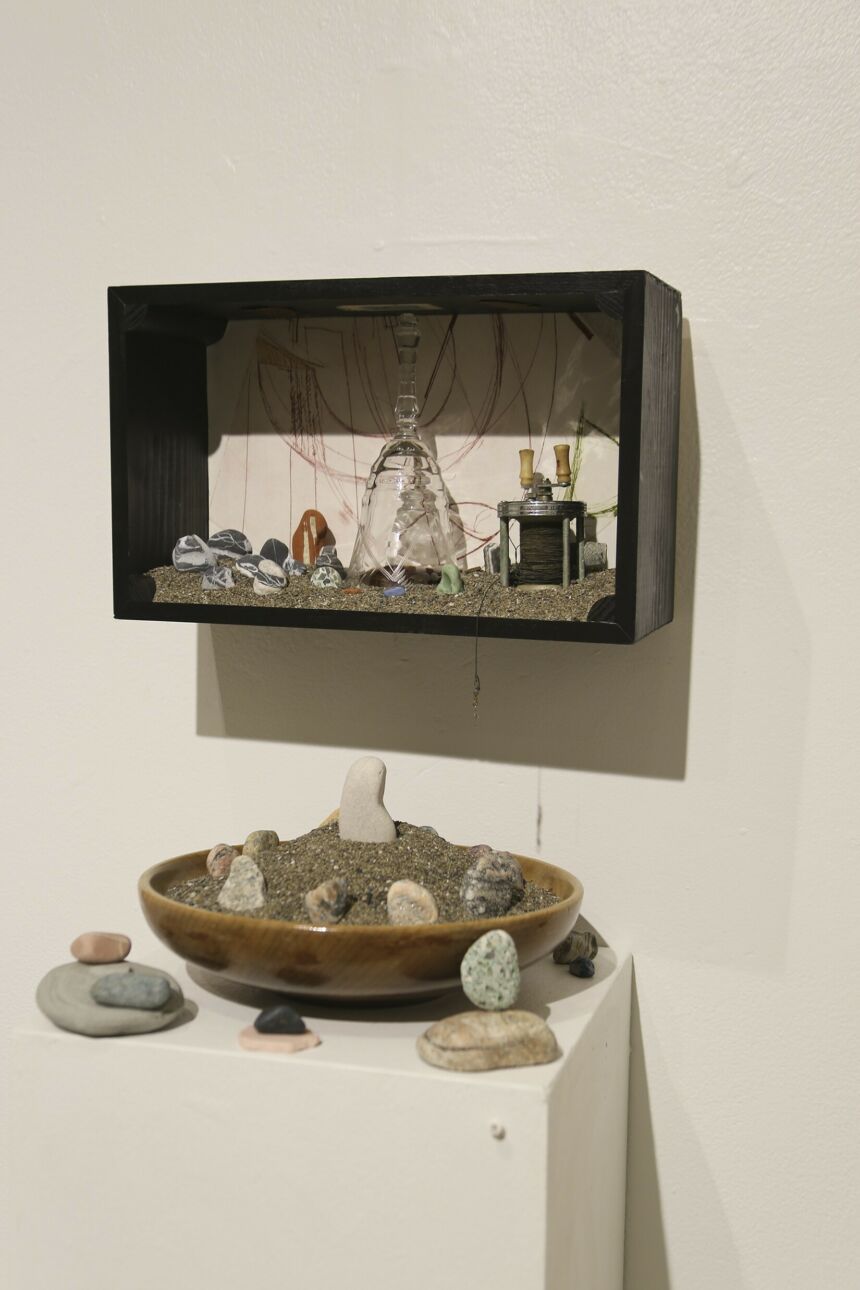 An upside-down glass goblet sits inside a wood shadow box with rocks, sand, and a fishing reel. Underneath sits a wooden bowl filled with sand, adorned with rocks in the sand and around the bowl.