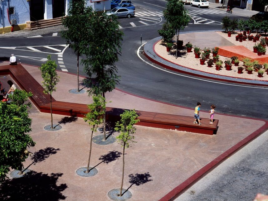 City area featuring a traffic circle, benches on which people are sitting and playing, trees, and planted shrubs with parked cars in the distance.