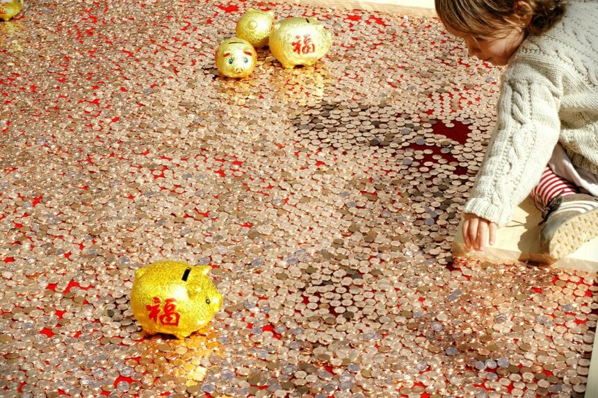 A young girl kneels on a floor covered in gold or copper coins and reaches to her left with god piggie banks with red script sit in the foreground and the background.