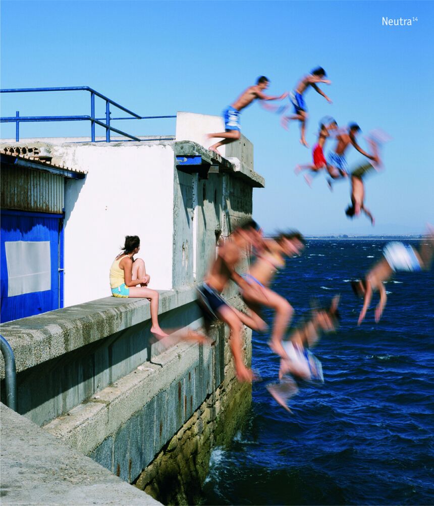 A group of nine young people jump from a barge into a body of water as a lone young person sits and looks on.