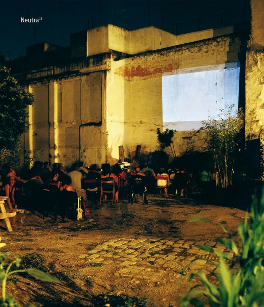 An exterior wall of a building at night with a screen projected on the side. Chairs with people sitting on them are facing the projected screen.