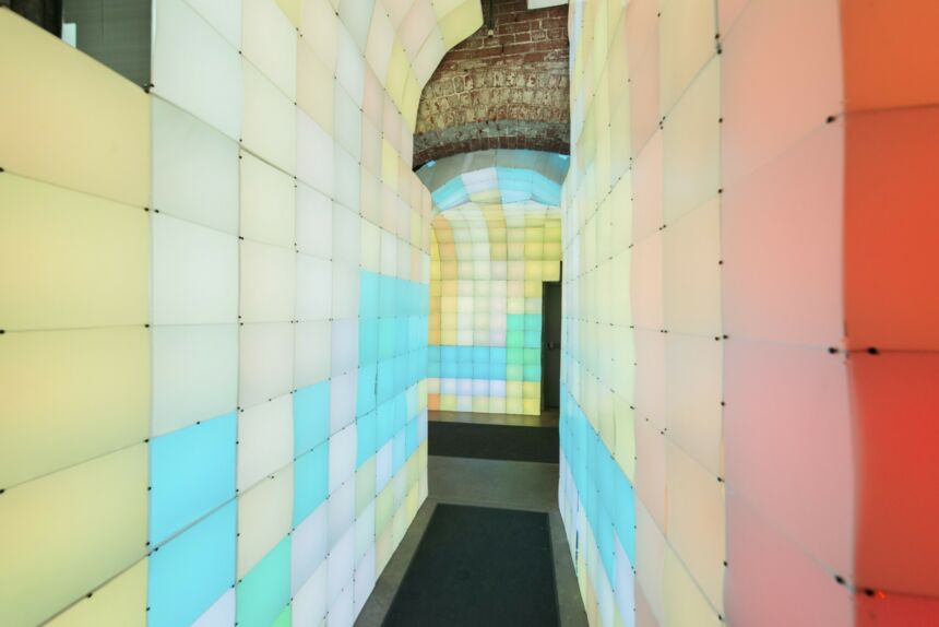 Hallway flanked by walls of light boxes illuminated in pastel green, yellow, pink, cerulean blue, white, and red.