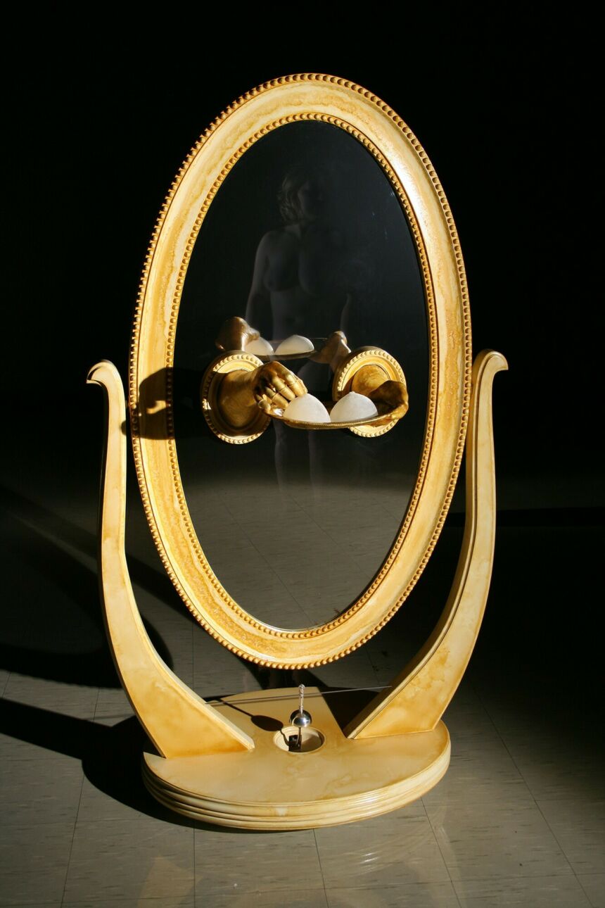 Gold disembodied hands protrude from an oval wood-framed floor mirror, holding a gold platter of white breast forms.