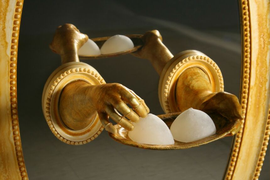 Close-up detail of gold disembodied hands protrude from an oval wood-framed floor mirror, holding a gold platter of white breast forms.
