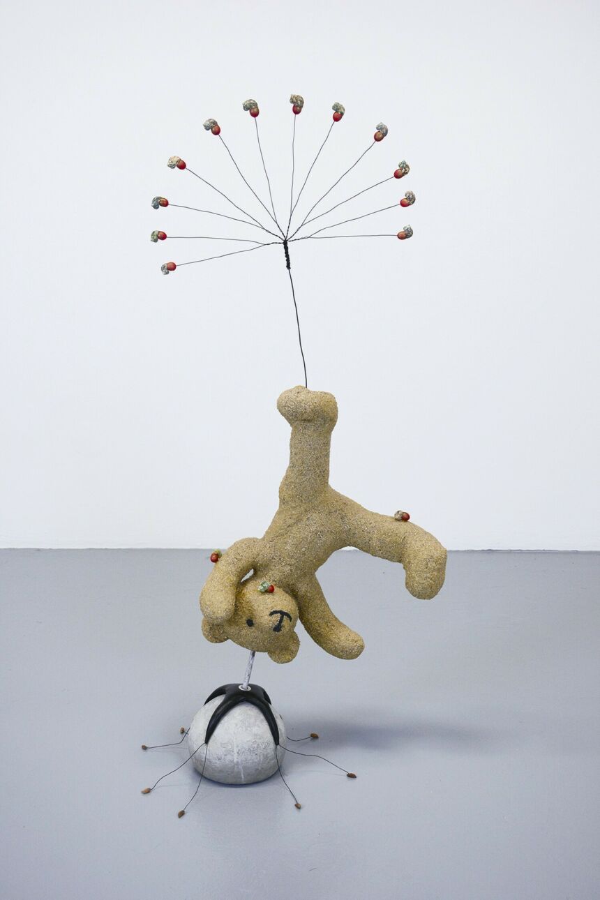 Sculpture of a teddy bear positioned in mid-cartwheel position, balanced on a white ball. A fan of decorative wires fan out from its left foot.