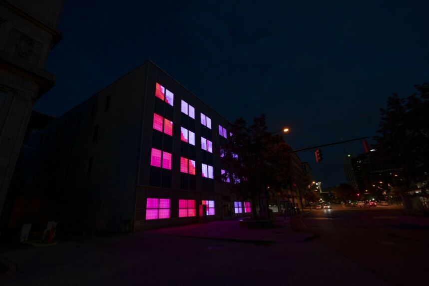 Exterior shot of warehouse at night, its windows are illuminated by pink, red, and purple light boxes.