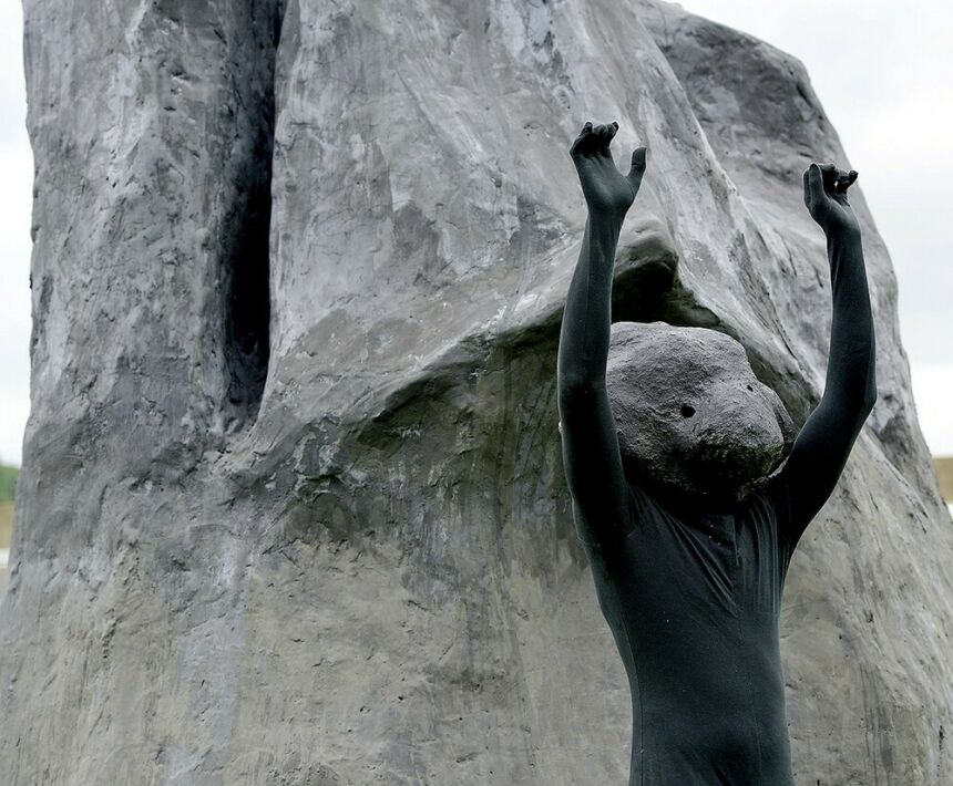 Artist Rudy Shepherd standing with arms raised front of his rock sculpture while wearing a black body suit and a faux rock "mask"