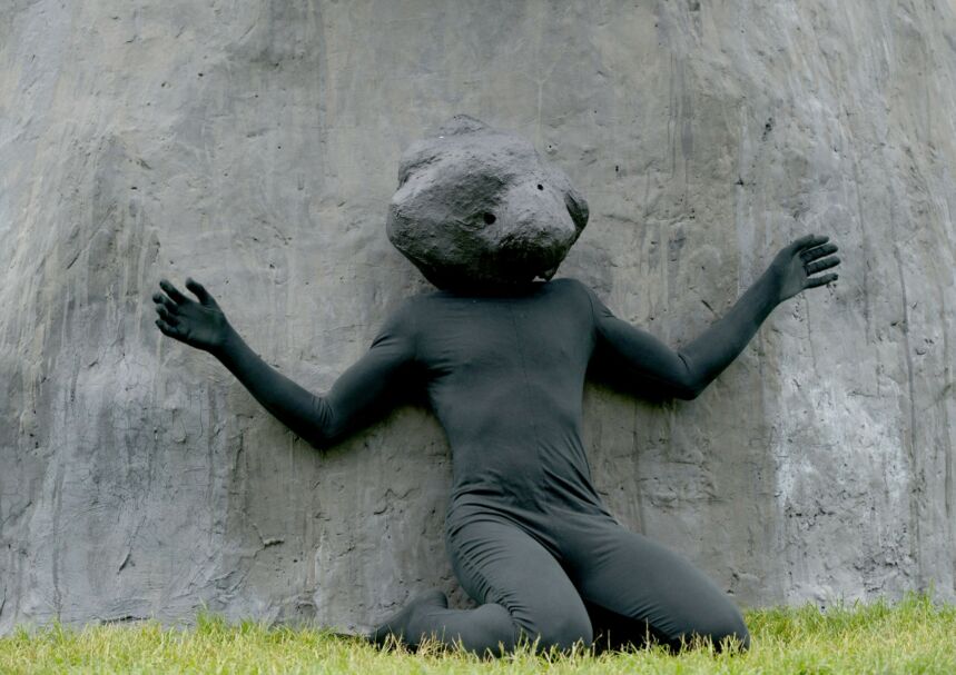 Artist Rudy Shepherd kneeling, arms spread in front of his rock sculpture while wearing a black body suit and a rock "mask"