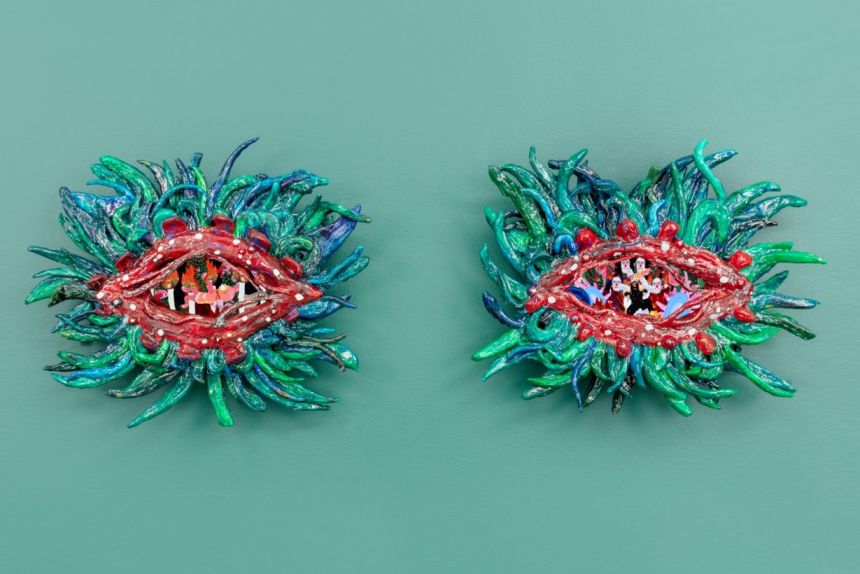 A pair of clay sculptures with eye-shaped apertures into performance stages featuring dancing actors. The red eye-lids are surrounded with green and blue floral lash-leaves.