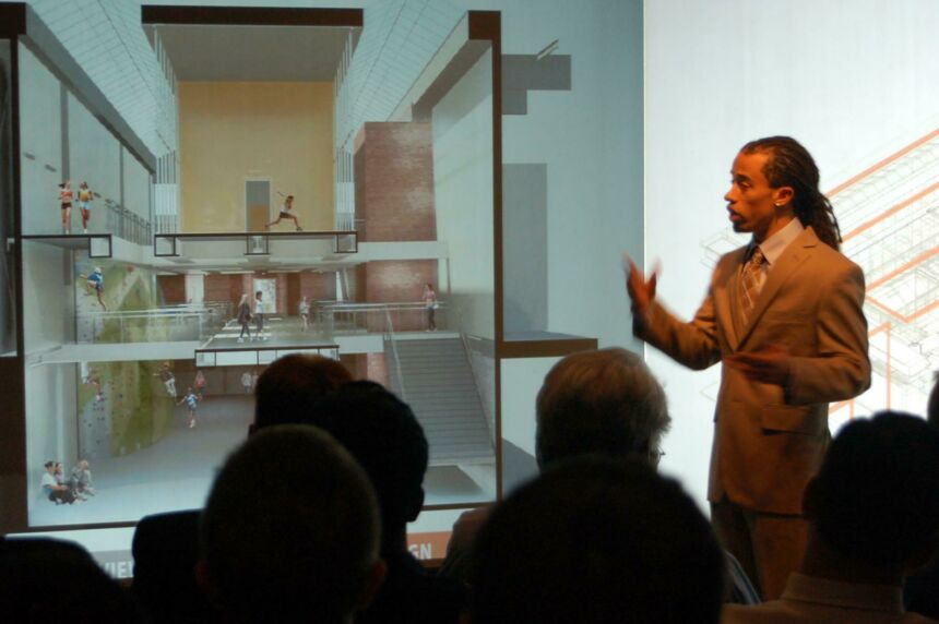 Photo of a presenter giving a lecture to an audience in front of a projection of an interior space design.