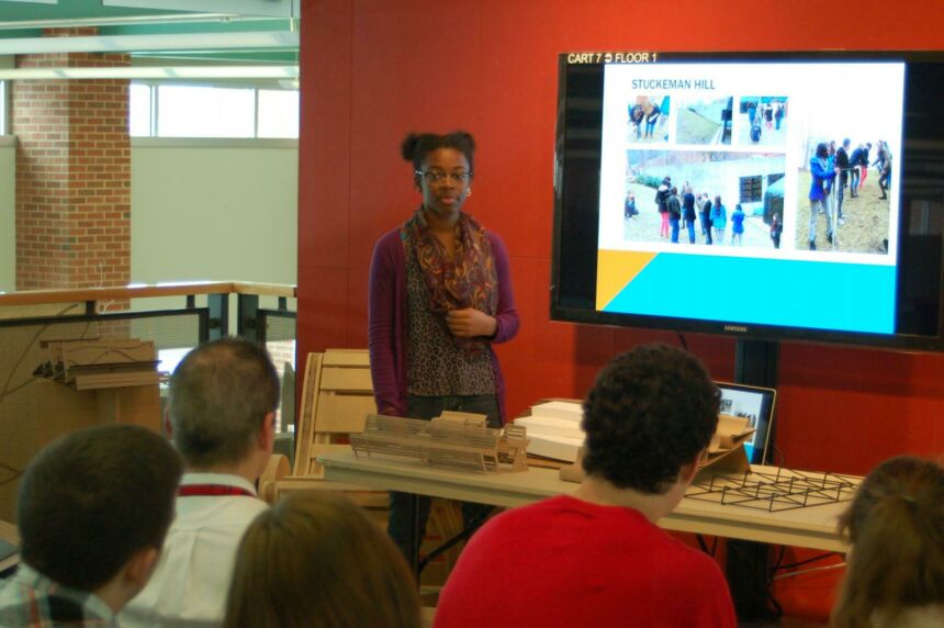 Photo of female student giving a presentation next to a monitor in front of her class.