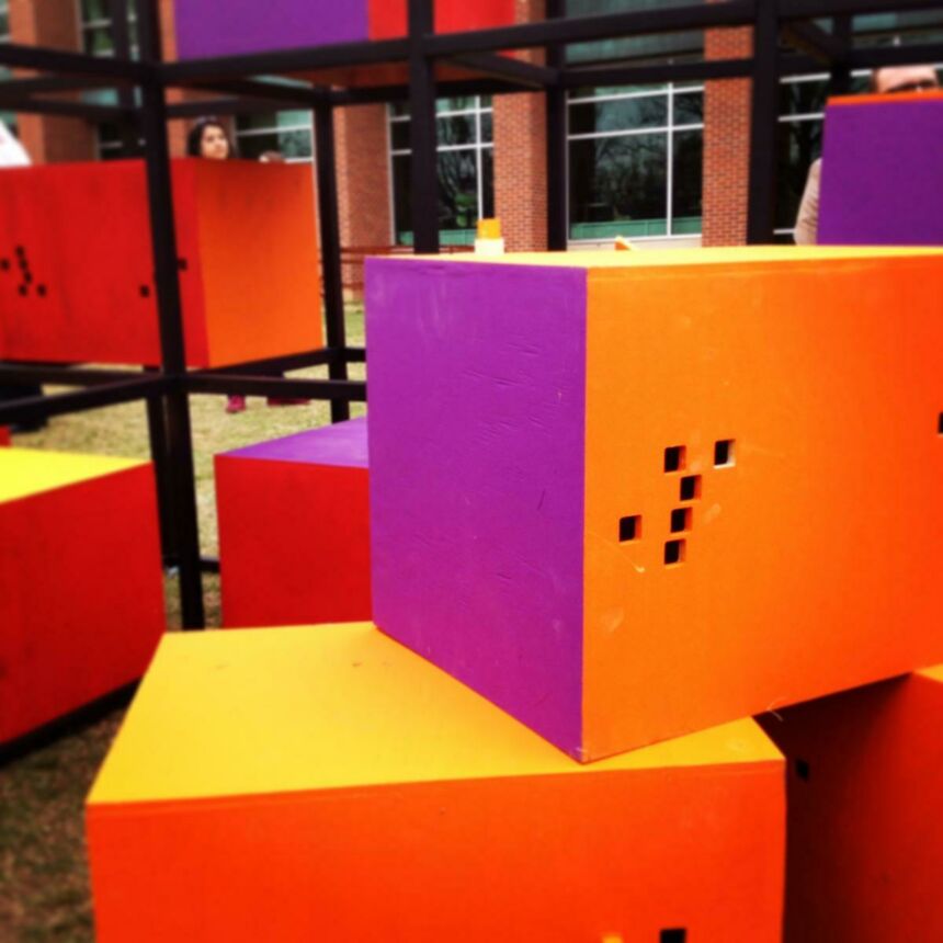Large wooden boxes painted red, orange, yellow, and orchid purple are stacked on one another, as well as on a black metal shelving unit in the background.