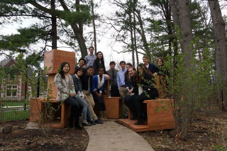 Group photo of Cathy Braasch and her students smiling at the camera while sitting on large wooden cubes among some trees on campus.