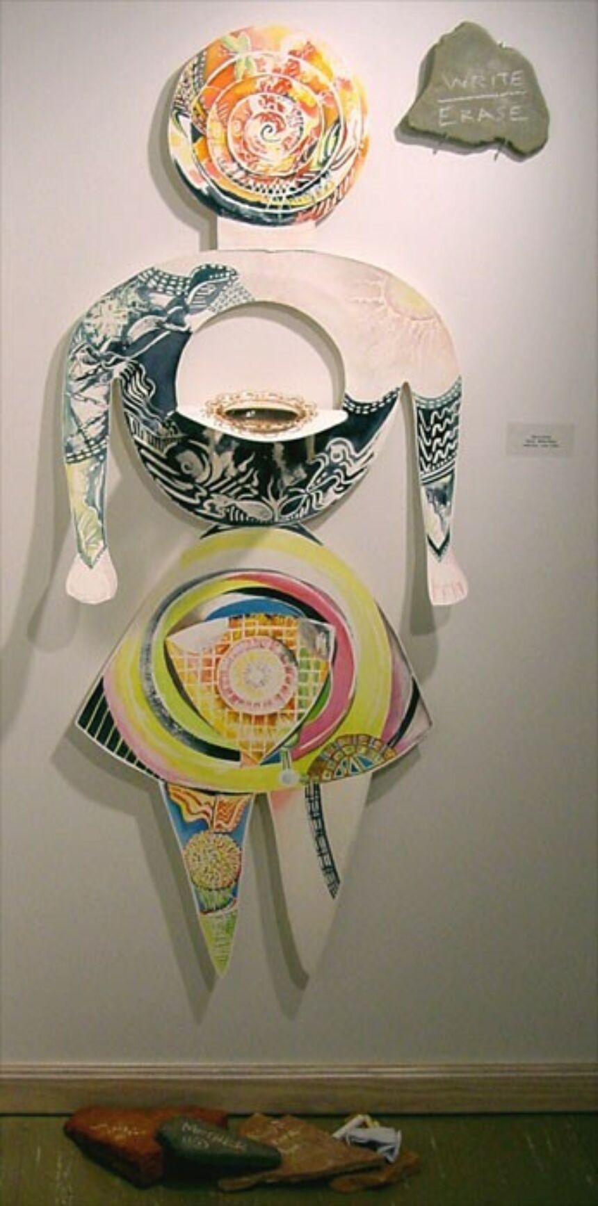 Wall installation of a vibrant watercolor piece, cut into an outline of a woman. A bowl sits on a flap cut out from the chest area. The title of the piece, "Write, Erase" is written on a flat stone affixed to the wall adjacent to the installation.