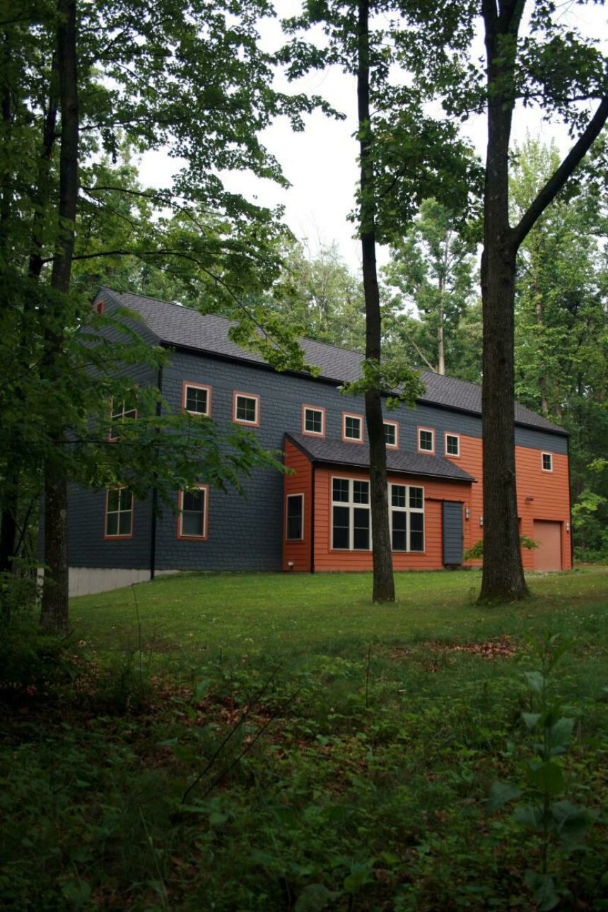 Exterior shot of a large, color-blocked grey and clay-red house surrounded by trees.