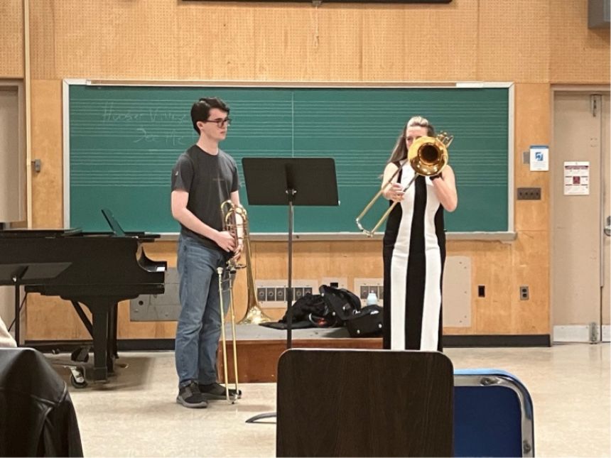 Woman in bold striped white and black suit playing a trombone in a classroom with a student standing close, paying attention.