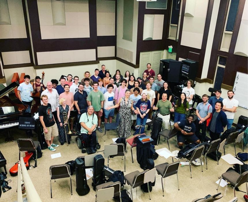 Overhead view of a large group of musicians clustered together for a group photo in a practice room littered with instruments, cases, stands and seats.