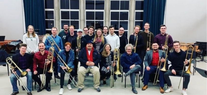 Three rows of trombonists sitting and standing for an informal group photo in a practice room.