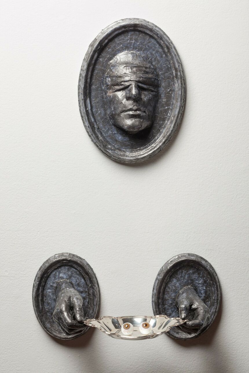Wall-mounted installation of a grey, blindfolded face and hands protrude from matching frames. The hands are holding a silver platter with two glass eyes.